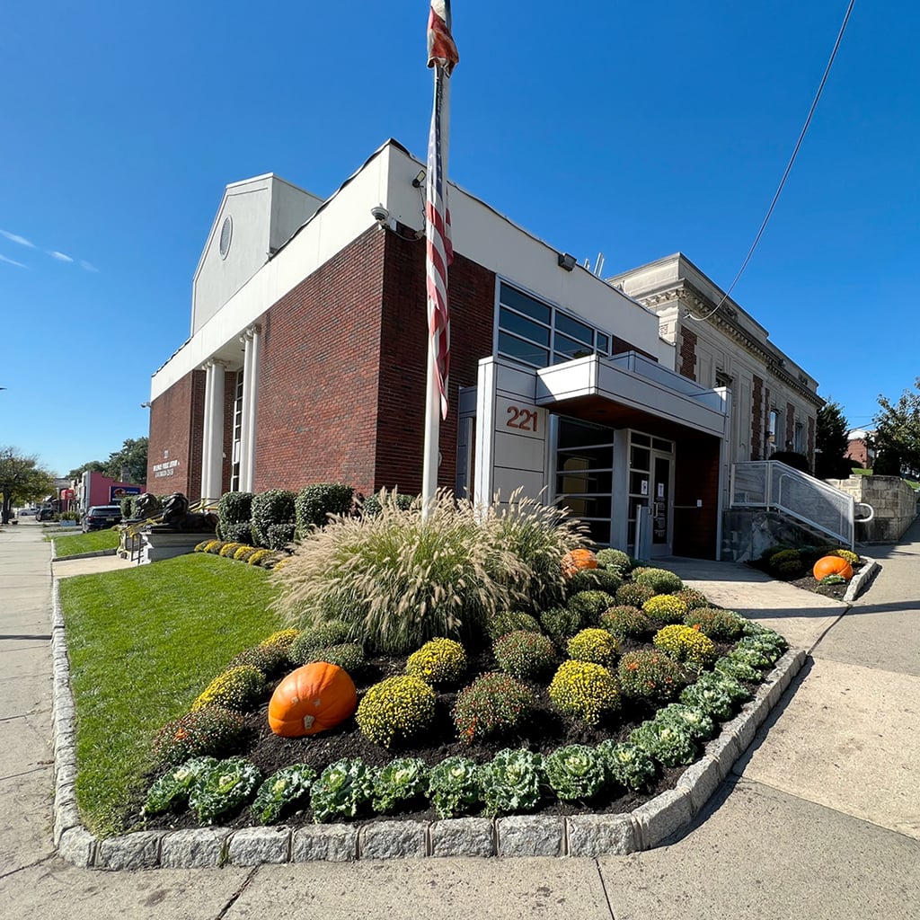 Exterior street view of the Belleville Public Library building, showing a brick facade, front entrance with address number 221, an American flag on a pole, and landscaped beds with pumpkins and fall flowers along the sidewalk.